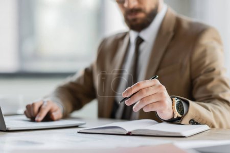 Teilaufnahme des Firmenchefs in beigem Blazer, Krawatte und Luxus-Armbanduhr, der mit Stift neben dem Notizbuch sitzt, während er am Arbeitsplatz im Büro am Laptop arbeitet