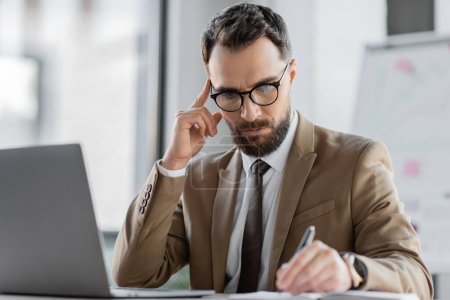 nachdenkliche und konzentrierte bärtige Manager in trendiger Business-Kleidung berühren Kopf beim Schreiben der Planung in Notizbuch in der Nähe Laptop auf dem Schreibtisch im Büro