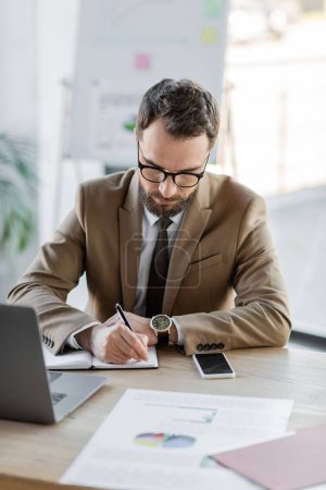 Photo for Successful entrepreneur in trendy blazer, eyeglasses and luxury wristwatch writing in notebook next to laptop, smartphone with blank screen and documents with charts on desk in office - Royalty Free Image