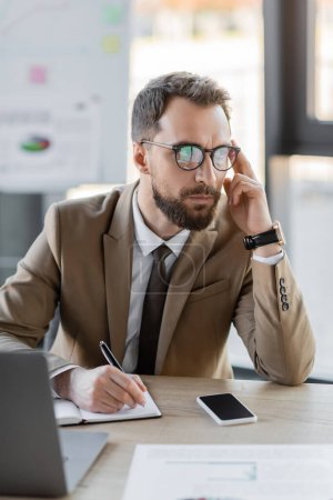 concentrated manager in business attire and eyeglasses touching head and writing in notebook near laptop, smartphone with blank screen and documents while thinking on business planning in office