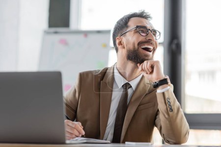 overjoyed and stylish businessman in beige blazer, tie and eyeglasses sitting at workplace with pen and notebook near laptop, laughing and looking away in office