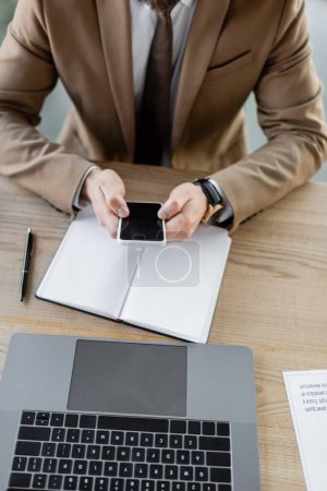 vista parcial del hombre de negocios en blazer beige que sostiene el teléfono inteligente con la pantalla en blanco cerca del cuaderno vacío, el ordenador portátil, la pluma y el documento en el escritorio en la oficina, vista de ángulo alto