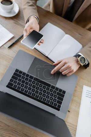 top view of cropped businessman in luxury wristwatch using touchpad of laptop while holding smartphone with blank screen near empty notebook, pen, documents and coffee cup on table