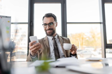 ambitious corporate manager in beige stylish blazer, eyeglasses and tie holding coffee cup and looking at mobile phone while smiling near notebook, saucer and computer on blurred foreground