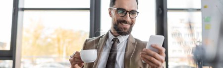 alegre hombre de negocios barbudo en blazer de moda, corbata y anteojos con taza de café y mirando el teléfono móvil en primer plano borroso en la oficina, pancarta