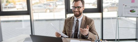 happy and successful entrepreneur in stylish eyeglasses, beige blazer and tie holding document and pointing with hand during video conference on laptop near notebooks in modern office, banner