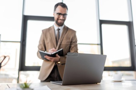 homme d'affaires barbu positif dans les lunettes, blazer beige et cravate debout près de l'ordinateur portable et tasse de café sur le bureau et l'écriture dans un carnet sur fond flou