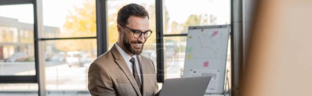 joyful bearded manager in eyeglasses, beige blazer and tie looking at laptop during video conference near flip chart with graphs and analytics in office, banner