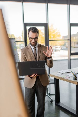 charismatic bearded businessman in eyeglasses and stylish formal wear smiling and waving hand during video conference on laptop next to desk with notebook and stationery in office, blurred foreground