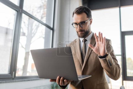 Photo for Serious bearded businessman in trendy eyeglasses, beige blazer and tie holding laptop and waving hand while having video conference near blurred windows in office - Royalty Free Image