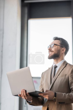 glücklicher und zufriedener Unternehmensmanager in Brille und modischer Businesskleidung wie beigem Blazer und Krawatte, der mit Laptop dasteht und im Büro wegschaut