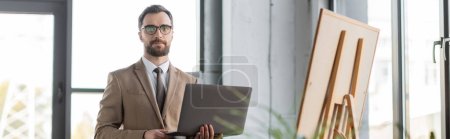 entrepreneur professionnel confiant en blazer beige, lunettes et cravate tenant ordinateur portable et regardant la caméra tout en se tenant près de flip chart dans le bureau contemporain, bannière