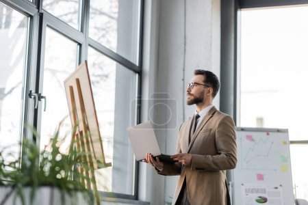 confident and fashionable bearded businessman with laptop looking away through windows near flip charts with infographics and blurred plants in contemporary office