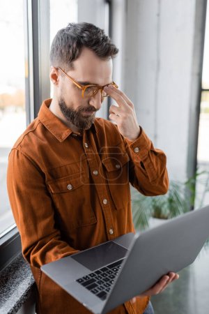 hombre de negocios barbudo concentrado en camisa de moda ajustando gafas con estilo y mirando a la computadora portátil borrosa mientras piensa cerca de la ventana en la oficina contemporánea
