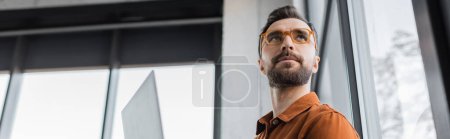 Photo for Low angle view of charismatic and brunette businessman with beard, wearing fashionable eyeglasses and shirt, looking away and thinking in office, banner, corporate lifestyle - Royalty Free Image