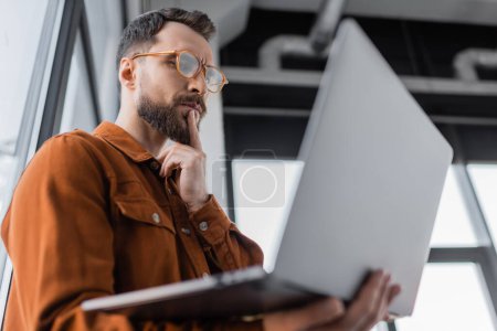 low angle view of serious bearded businessman in trendy eyeglasses and shirt looking at laptop and thinking while standing in office on blurred foreground, corporate lifestyle 