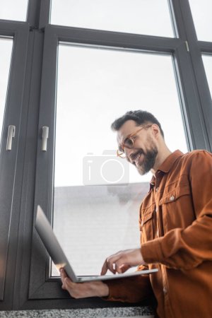 low angle view of successful and bearded businessman in eyeglasses and stylish shirt standing near window in modern office and smiling while using laptop, corporate lifestyle 