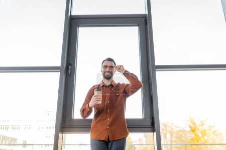 low angle view of cheerful accomplished businessman in fashionable shirt standing with coffee to go near large office windows, adjusting eyeglasses and looking at camera 