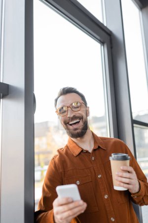 überglücklicher Geschäftsmann in stylischem Hemd und trendiger Brille, der mit Smartphone und Coffee to go neben dem Fenster im Büro steht und mit geschlossenen Augen lacht