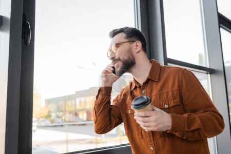 pleased bearded businessman in fashionable eyeglasses and stylish shirt holding paper cup during conversation on smartphone near window in contemporary office