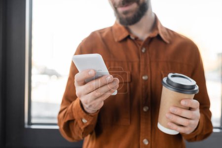 Teilbild des bärtigen Geschäftsmannes im schicken Hemd mit Handy, während er mit Kaffee in Pappbecher am Fenster im Büro steht, verschwommener Hintergrund