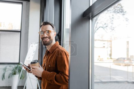 good looking and joyful businessman in stylish shirt and eyeglasses standing with disposable cup and mobile phone near windows in office and looking at camera