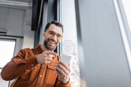 low angle view of excited entrepreneur with beard and eyeglasses, wearing stylish shirt, pointing with finger at mobile phone during video call in modern office 