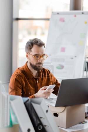 fokussierter bärtiger Geschäftsmann in trendiger Brille und Hemd sitzt mit Handy neben Ordnern, Laptop auf Karton und Flipchart auf verschwommenem Hintergrund im Büro