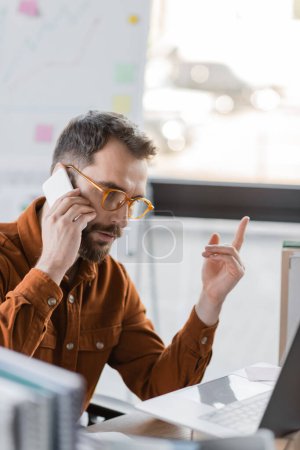 Foto de Hombre de negocios barbudo enfocado en anteojos y camisa hablando en el teléfono inteligente y mostrando el gesto de atención cerca de la computadora portátil y los cuadernos borrosos en el lugar de trabajo en la oficina - Imagen libre de derechos