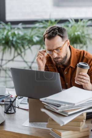 serious bearded businessman with paper cup adjusting eyeglasses and thinking near laptop on carton box, smartphone, pile of books and notebooks and pen holder with stationery on desk in office