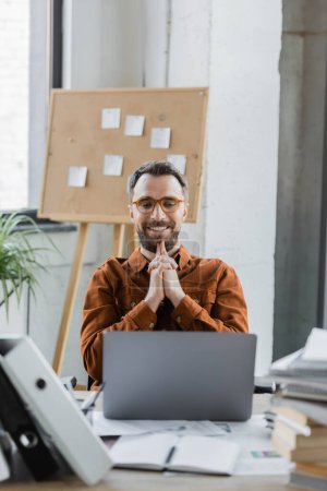satisfied bearded businessman in trendy shirt and eyeglasses looking at laptop near blurred folders, notebooks, documents and flip chart with sticky notes on background in office