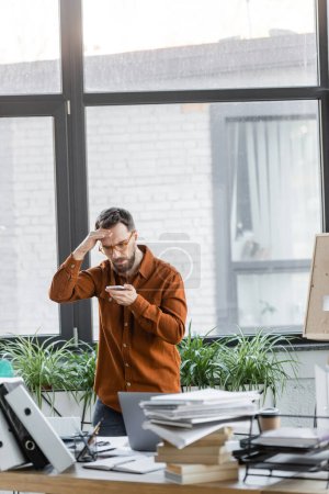 worried businessman in eyeglasses looking at mobile phone and holding hand near head next to work desk with laptop, notebooks, folders, books, paper cup and decorative plants in modern office