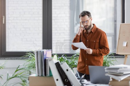 thoughtful and busy businessman in eyeglasses looking at document while standing next to work desk with laptop, plenty of books, notebooks, folders and corkboard with sticky note on background