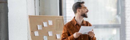 accomplished and happy bearded businessman in eyeglasses and stylish shirt looking away near corkboard with attached sticky notes in modern office, banner