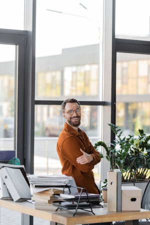happy bearded businessman in eyeglasses and shirt smiling at camera while standing with crossed arms next to windows and workplace with pile of books, notebooks, folders, carton box and paper cup 