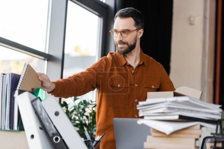 hombre de negocios barbudo positivo en camisa y anteojos que sostiene la caja de cartón y toma el cuaderno en el lugar de trabajo con el ordenador portátil, un montón de cuadernos y carpetas en la oficina