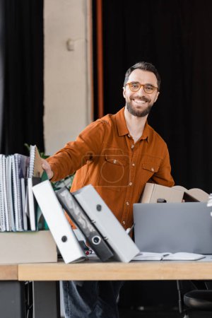 fröhlicher Manager in schickem Hemd und Brille mit Kartonschachtel und lächelnd in die Kamera neben dem Arbeitsplatz mit Notizbüchern, Ordnern und Laptop im modernen Büro