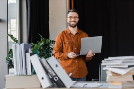 happy successful businessman in stylish eyeglasses and shirt standing with laptop and looking at camera near pile of books, notebooks and folders on blurred foreground in office