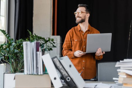 positive and successful entrepreneur in eyeglasses and shirt standing with laptop and looking away near blurred pile of books, notebooks and folders on work desk in office