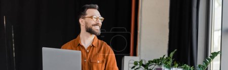 overjoyed and bearded executive manager in stylish eyeglasses and shirt smiling and looking away near laptop and decorative plants in modern office, banner