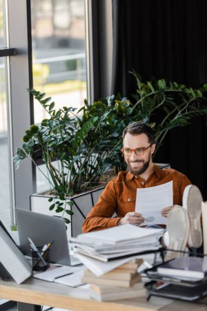 Foto de Hombre de negocios carismático y feliz en gafas con documento mientras está sentado con las piernas en el escritorio cerca de la computadora portátil, pila de cuadernos y plantas decorativas en la oficina - Imagen libre de derechos