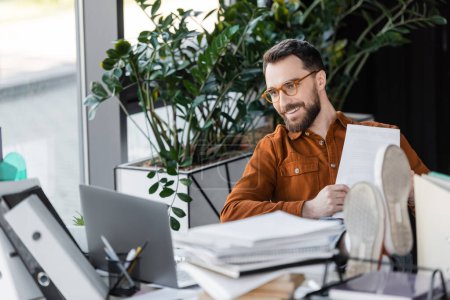 Foto de Empresario barbudo sonriente en gafas de moda y camisa sosteniendo papel y mirando a la computadora portátil mientras está sentado con las piernas en el escritorio cerca de los cuadernos y carpetas borrosas en la oficina - Imagen libre de derechos