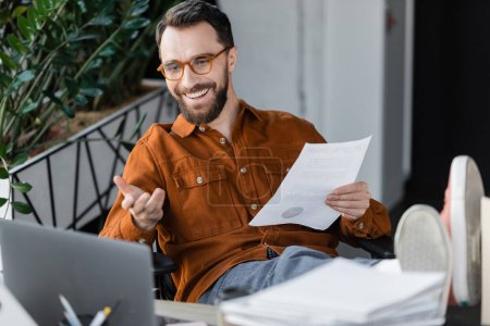 cheerful businessman in trendy eyeglasses and shirt holding document while sitting with legs on desk and pointing with hand during video call on laptop near papers on blurred foreground