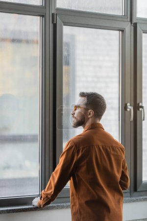 Photo for Brunette and bearded businessman in stylish eyeglasses and fashionable shirt, confident and successful, standing at large window in office and looking away - Royalty Free Image