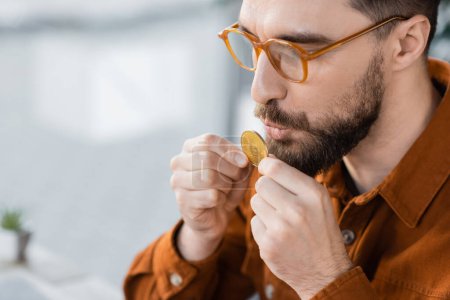 Photo for KYIV, UKRAINE - OCTOBER 18, 2022: bearded and successful businessman in fashionable eyeglasses and stylish shirt holding and kissing golden bitcoin on blurred background in contemporary office - Royalty Free Image