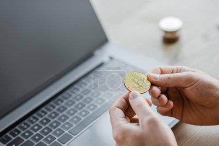 Photo for KYIV, UKRAINE - OCTOBER 18, 2022: cropped view of ambitious businessman holding golden bitcoin near laptop with blank screen on office desk, blurred background - Royalty Free Image