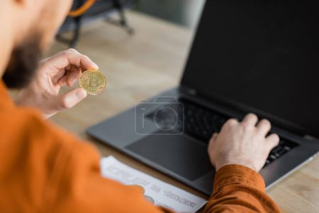 Photo for KYIV, UKRAINE - OCTOBER 18, 2022: partial view of blurred successful entrepreneur holding golden bitcoin and working on laptop with blank screen in office - Royalty Free Image