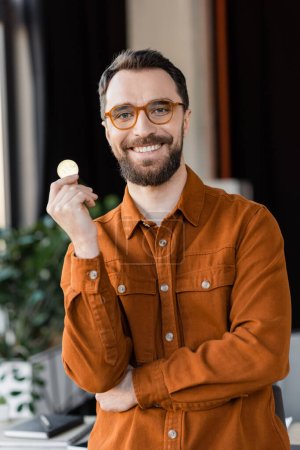 Photo for KYIV, UKRAINE - OCTOBER 18, 2022: charismatic and bearded businessman in trendy eyeglasses and shirt holding golden bitcoin and smiling at camera in modern office on blurred background - Royalty Free Image