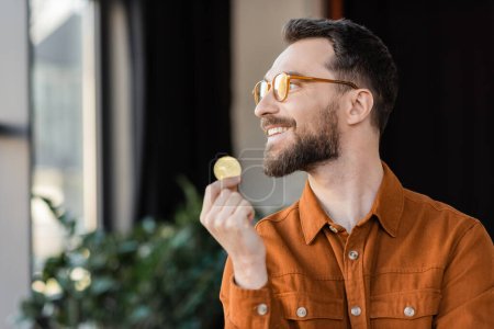 Photo for KYIV, UKRAINE - OCTOBER 18, 2022: joyful and accomplished businessman in stylish eyeglasses and shirt holding golden bitcoin, smiling and looking away in modern office on blurred background - Royalty Free Image