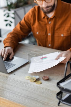 Photo for KYIV, UKRAINE - OCTOBER 18, 2022: partial view of businessman in eyeglasses and shirt using laptop and holding documents near silver and golden bitcoins on work desk in office - Royalty Free Image
